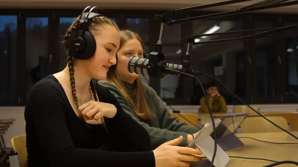 Zwei Schülerinnen nehmen mit Mikrofon und Kopfhörern einen Audiobeitrag auf.