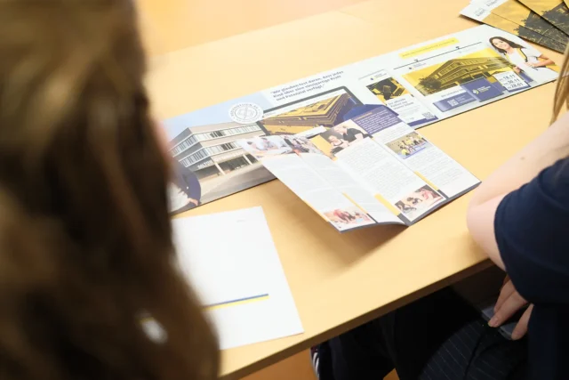 Schülerinnen betrachten gemeinsam einen gestalteten Flyer auf einem Tisch.