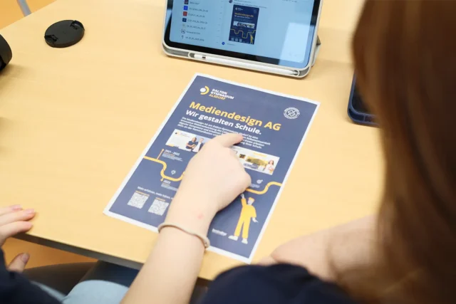 Schülerin zeigt auf eine gestaltete Broschüre, daneben liegen Tablet und Kamera.