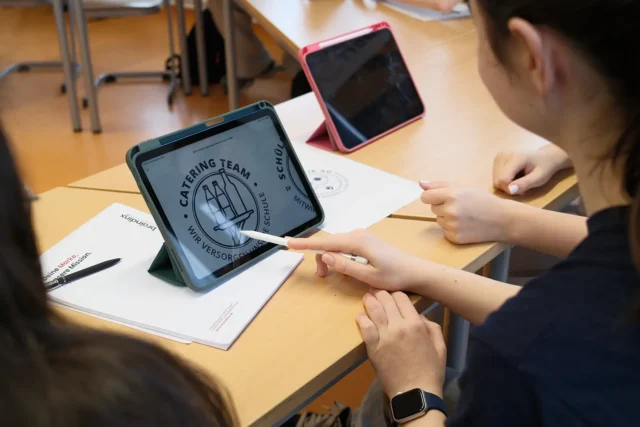 Schülerinnen arbeiten gemeinsam an einem Logo-Entwurf auf einem Tablet.