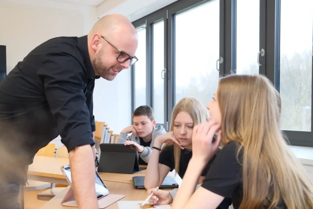 Lehrer unterstützt Schülerinnen bei der Arbeit an einem Designprojekt im Klassenraum.